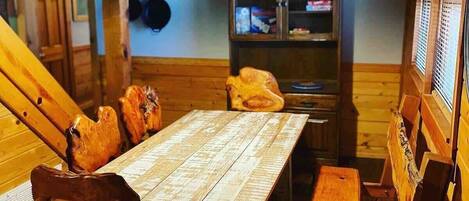 A rustic wooden dining room with a large table and chairs, a hanging light fixture, and a kitchen area with cabinets and a stove.