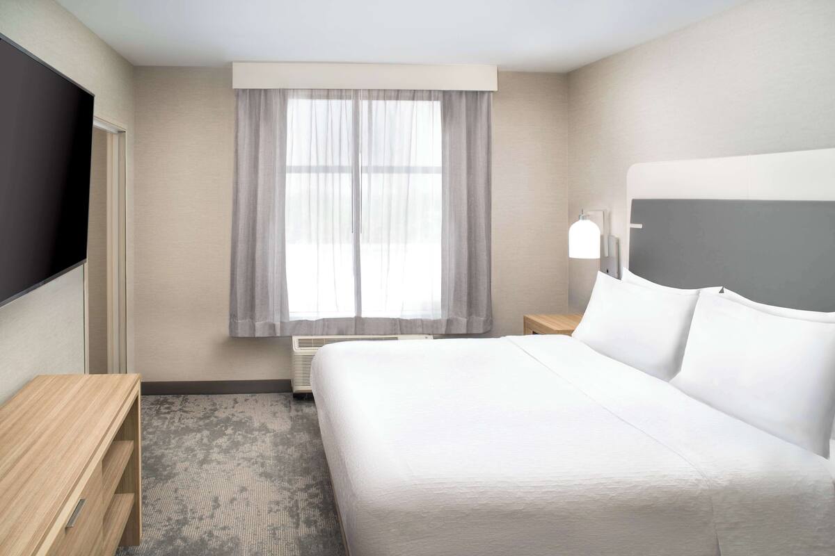 Habitación de hotel con una cama grande, una mesita de noche de madera, un televisor de pantalla plana, una ventana con cortinas translúcidas y una lámpara fijada a la pared.