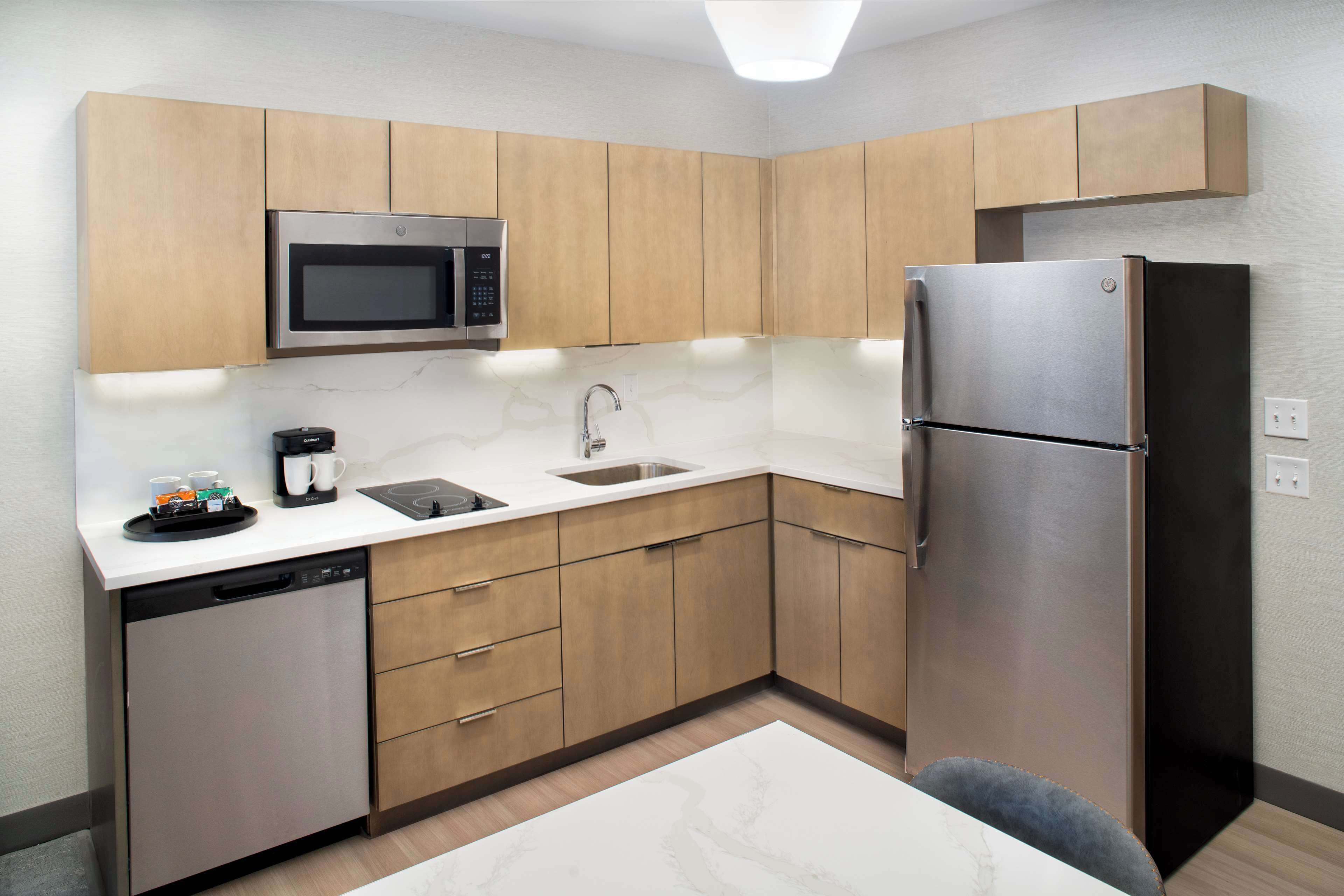 A compact kitchen with wooden cabinets, a stainless steel refrigerator, a microwave, a dishwasher, and a sink.
