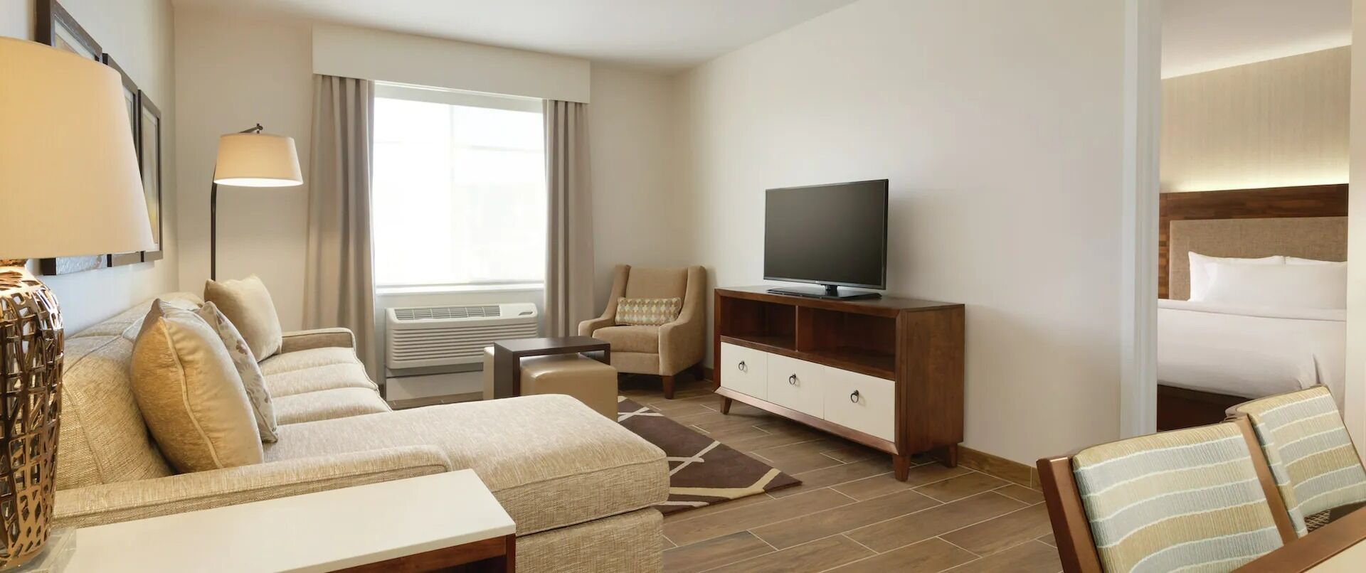 Una habitación de hotel con sofá, televisión sobre un mueble de madera, una silla, cama y ventana con cortinas.