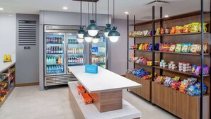 Un hall d’hôtel doté d’un mini-bar, d’un espace de libre-service pour les boissons, ainsi que d’étagères remplies d’en-cas et de produits de toilette.