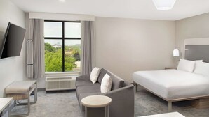 Une chambre d’hôtel moderne avec un lit, un canapé et une petite table. Un téléviseur est fixé au mur et il y a une fenêtre avec des rideaux.