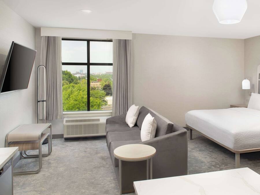 Una habitación de hotel moderna con cama, sofá y una pequeña mesa. Hay un televisor montado en la pared y una ventana con cortinas.