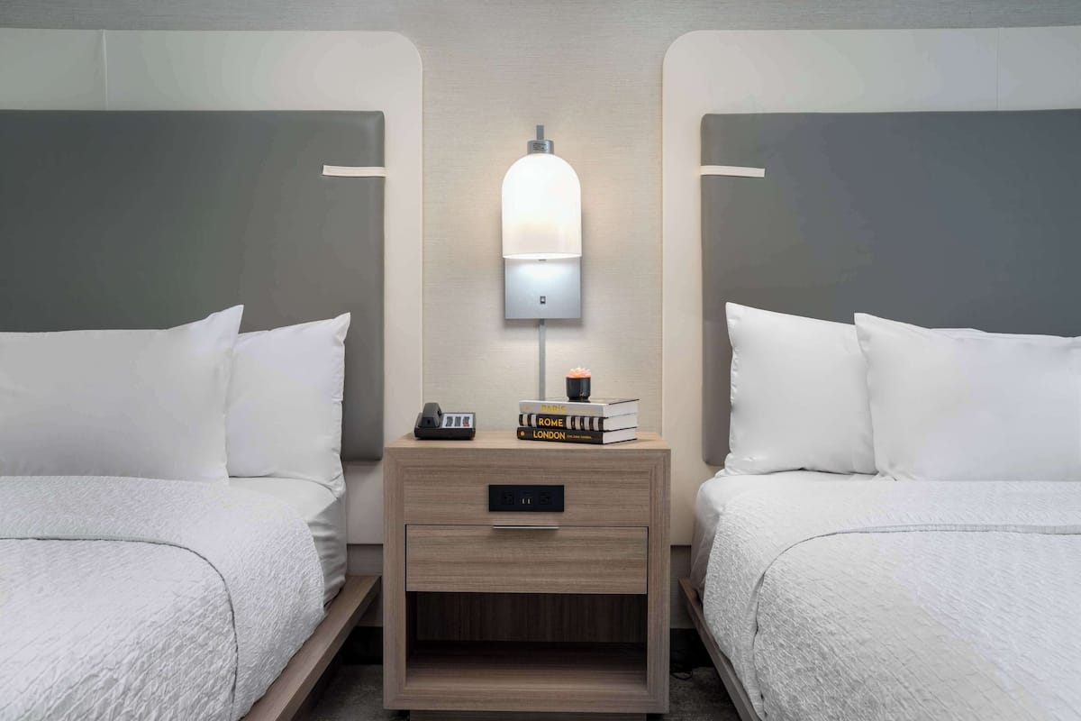 Habitación de hotel con dos camas, una mesita de noche con lámpara y un cajón pequeño.