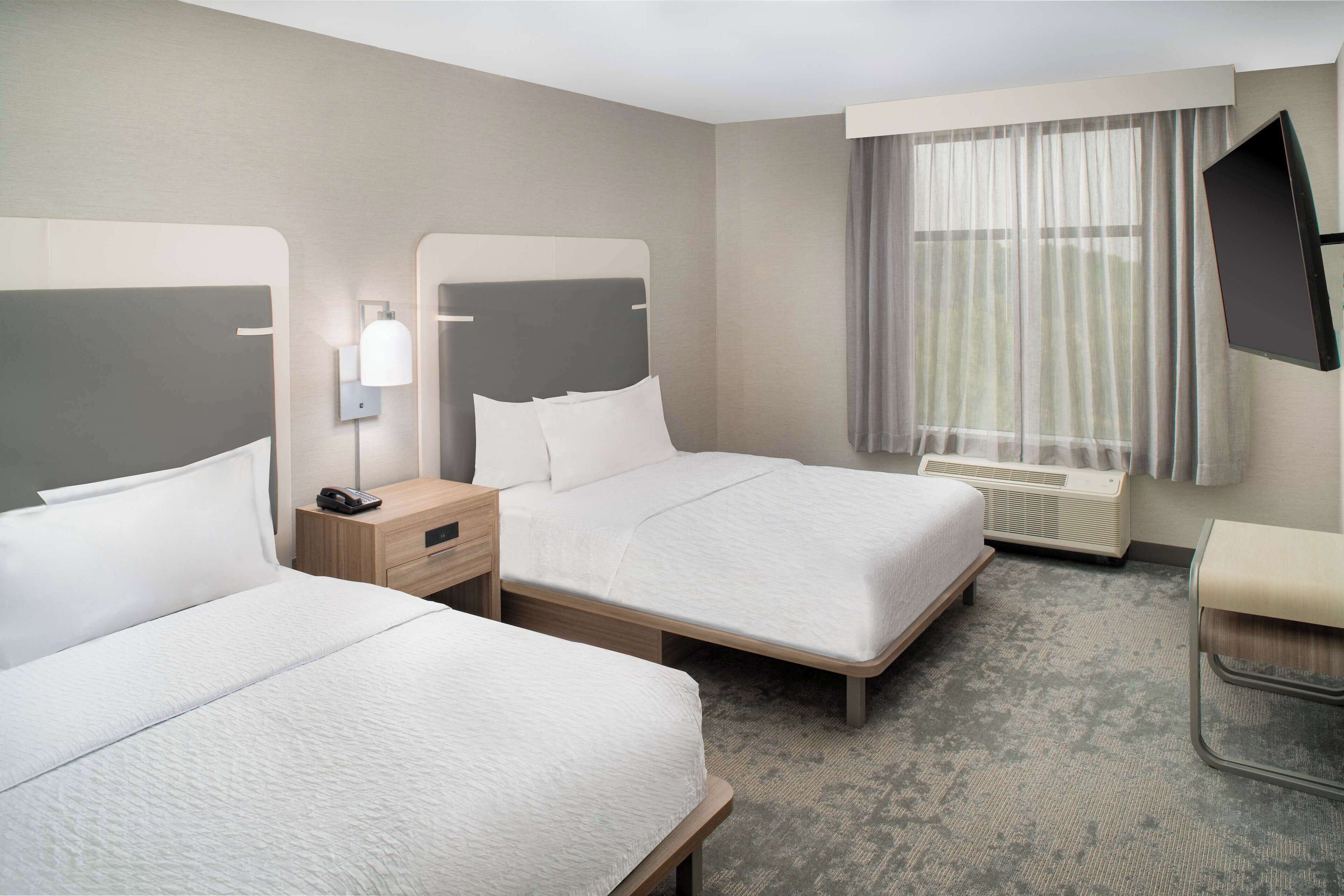A hotel room with two beds, a bedside table with a lamp, a TV mounted on the wall, and a window with curtains.