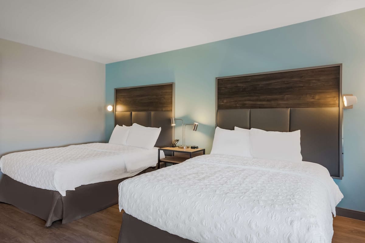 Une chambre d’hôtel avec deux lits, chacun doté d’une tête de lit en bois et de linge de lit blanc. Une table de chevet se trouve entre les lits, avec une lampe et un téléphone.