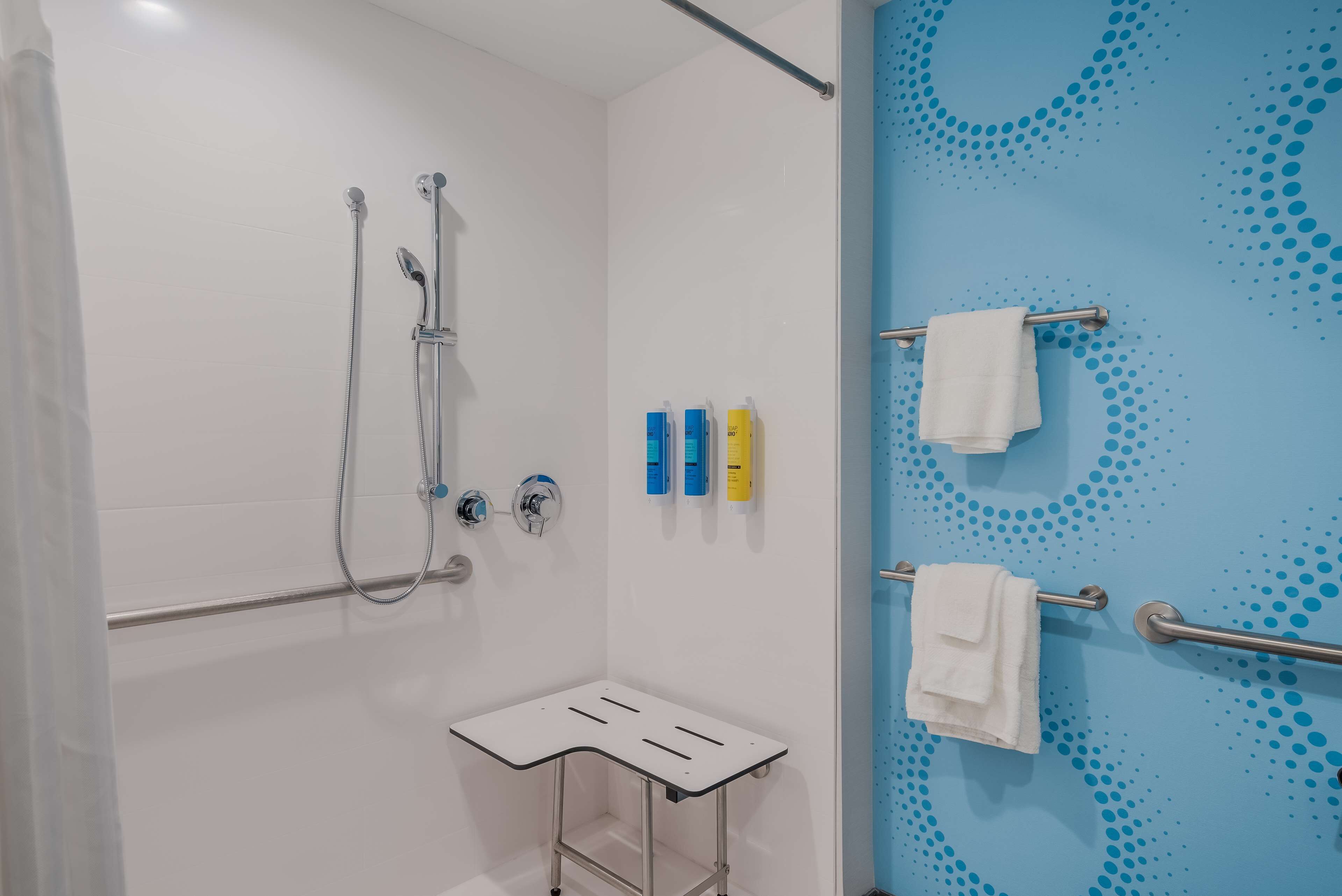 Un espace douche doté d’un receveur de douche blanc, d’un pommeau de douche à main et d’un robinet mural. Trois flacons de produits de toilette sont fixés au mur, et deux serviettes sont disposées sur des porte-serviettes.