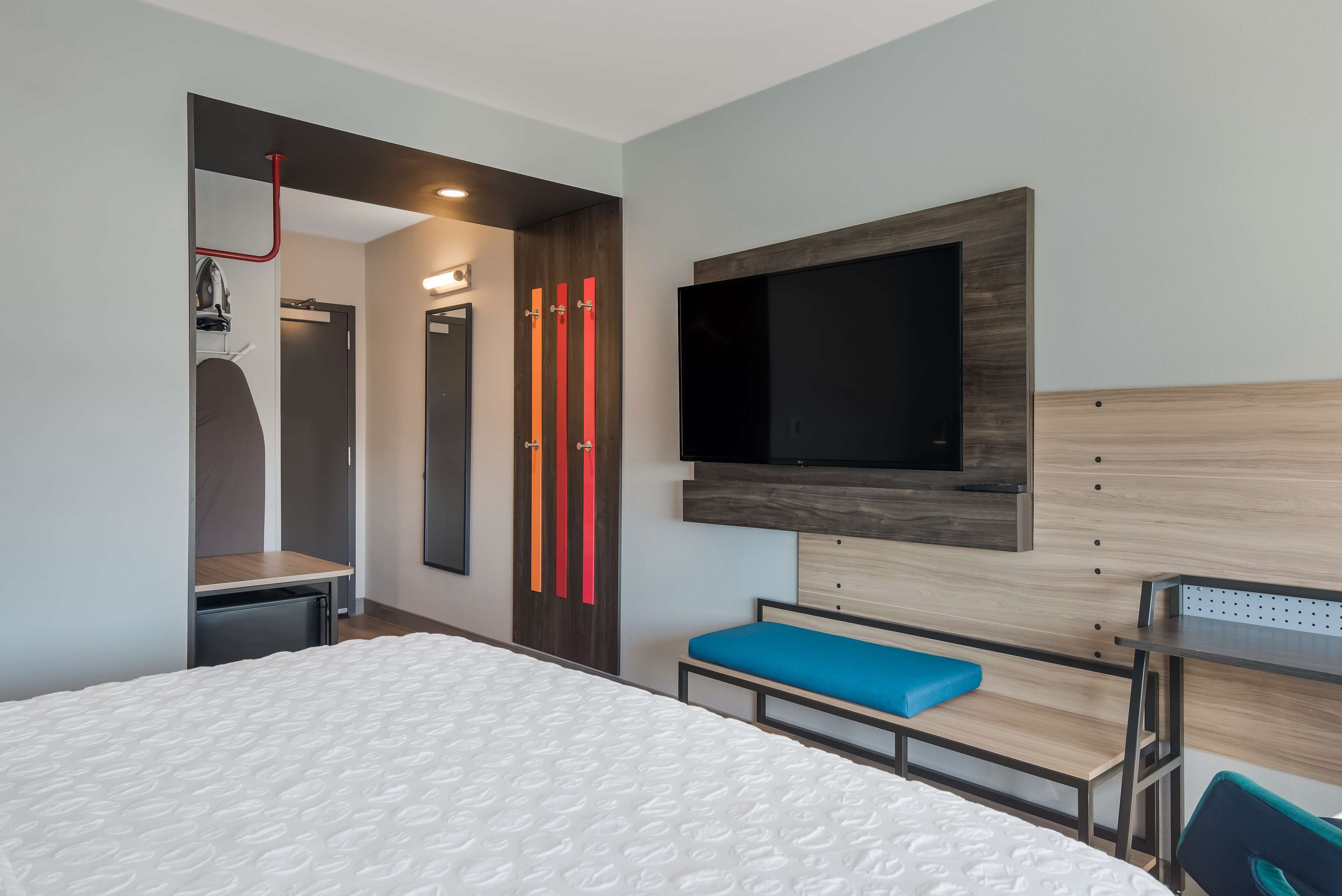 Habitación de hotel moderna con una cama grande, un escritorio con silla, un televisor montado en la pared y un banco con cojín azul.