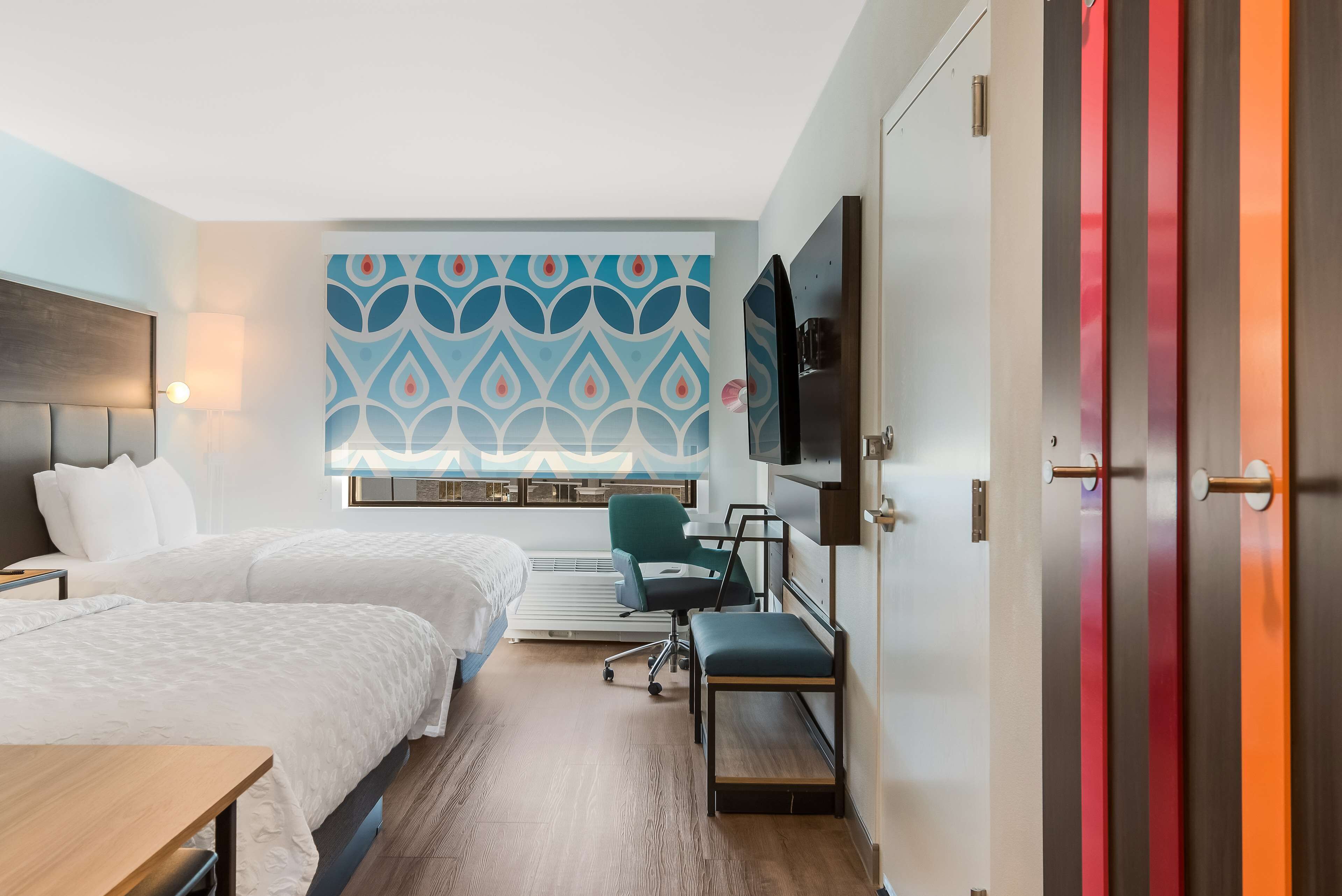 A hotel room with two beds, a desk, a chair, a TV, and a patterned wall.