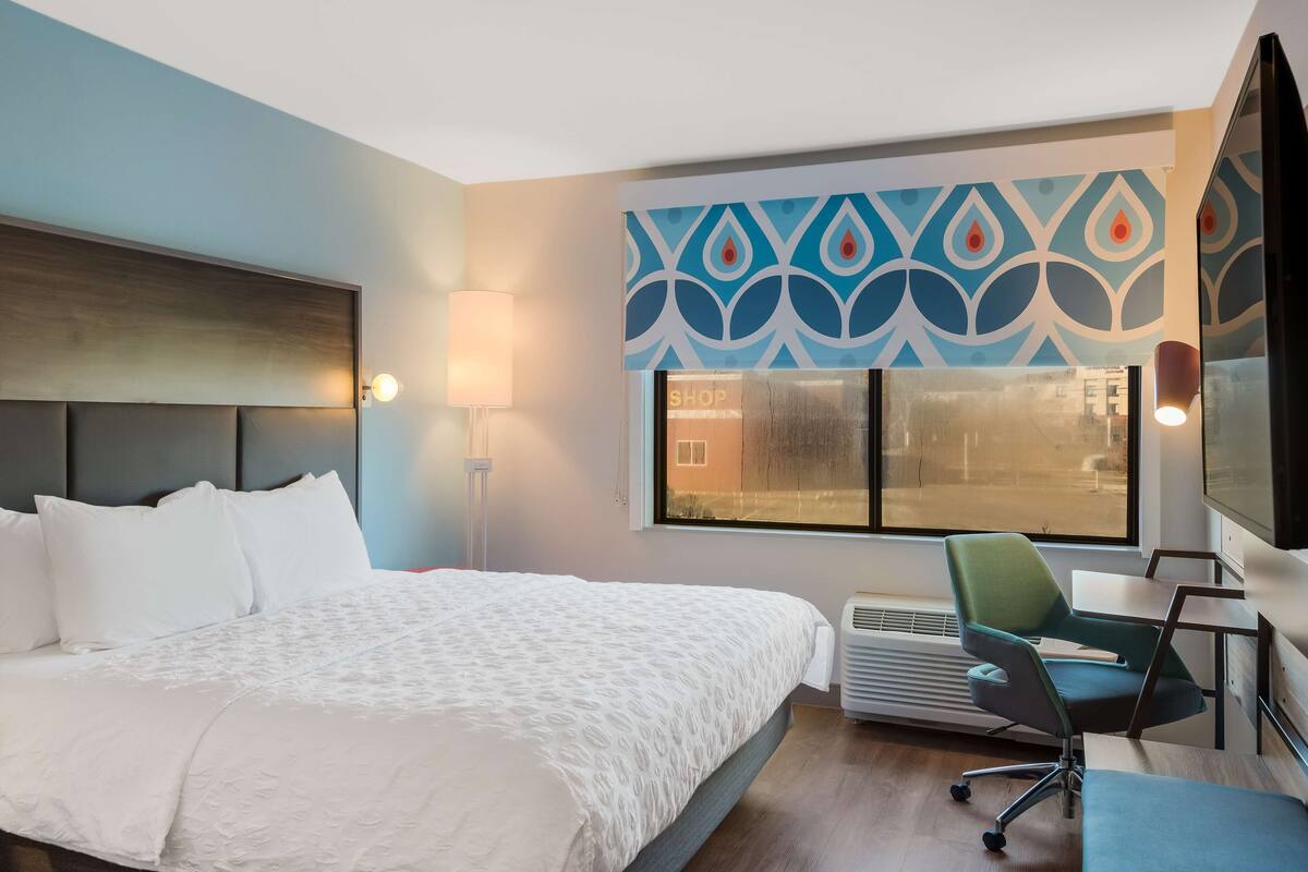 Une chambre d’hôtel avec un grand lit, un bureau avec une chaise, une fenêtre donnant sur un paysage agréable et un mur orné d’un motif décoratif.