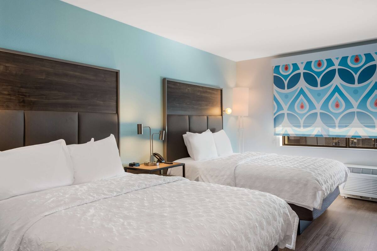 Une chambre d’hôtel avec deux lits, une table de chevet avec une lampe et une grande fenêtre ornée d’un rideau à motifs.