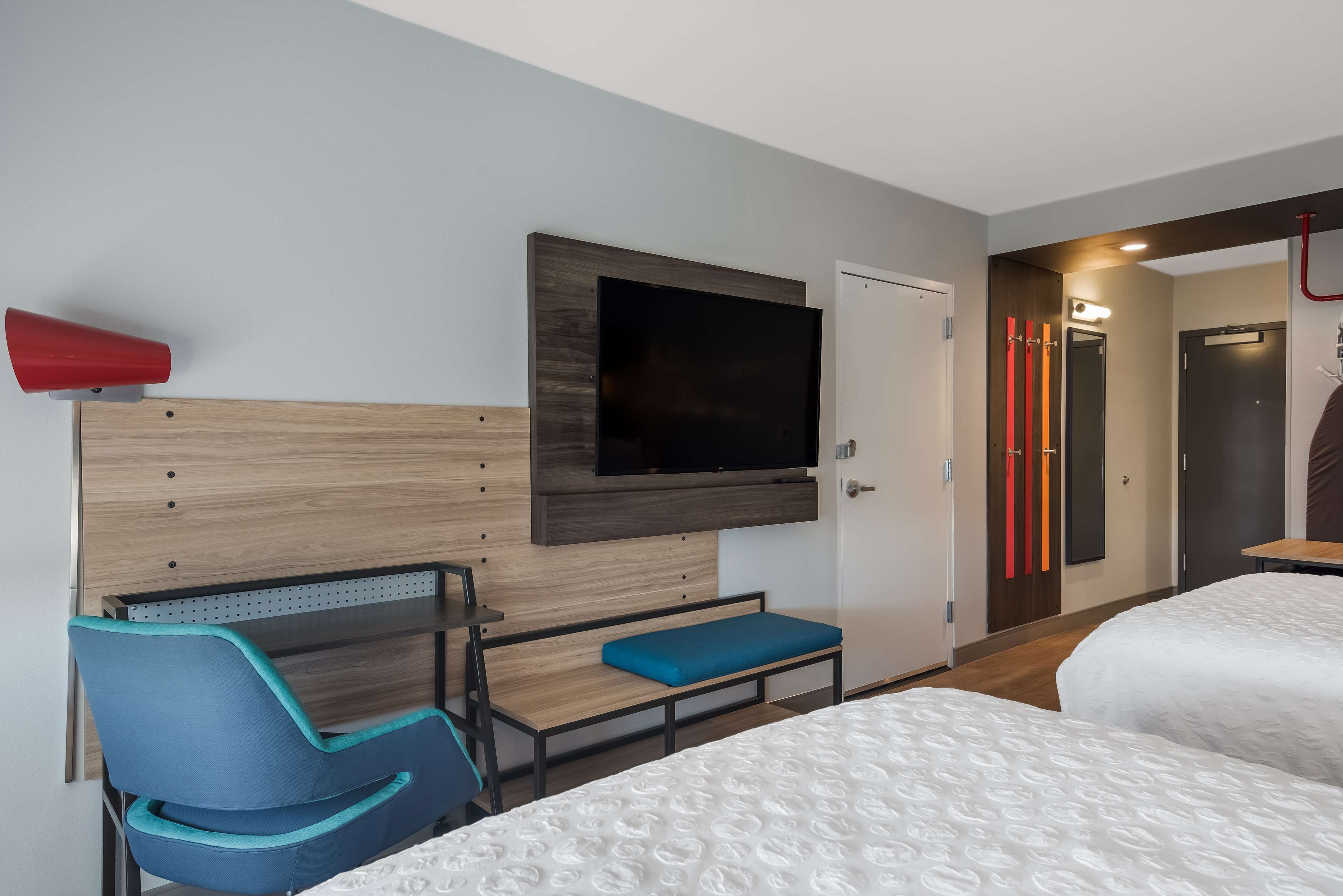 A hotel room with a bed, a seating area with a blue chair, a television mounted on the wall, and a door with a light above it.