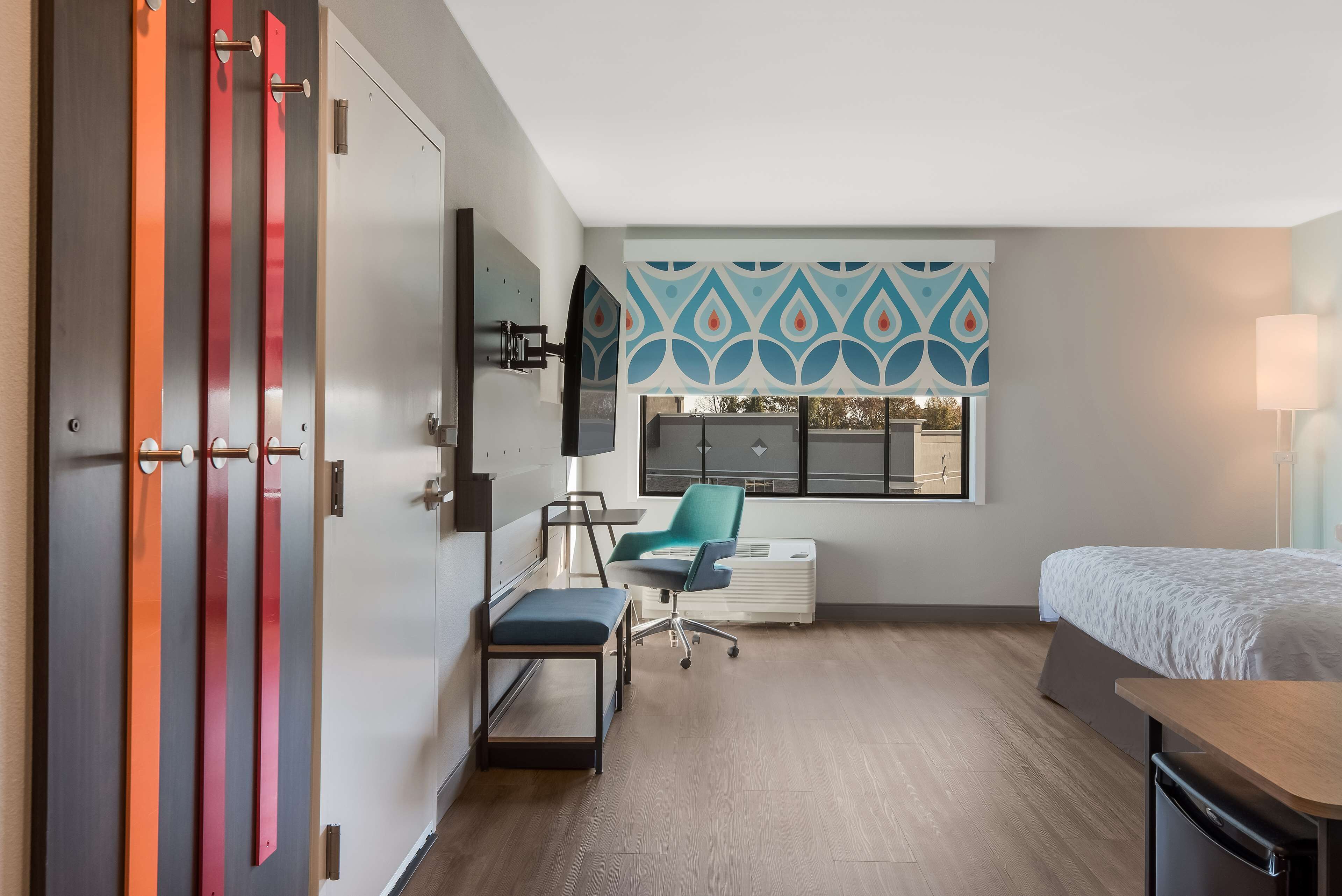 Habitación de hotel con cama, escritorio, silla, televisión y ventana con cortina estampada.
