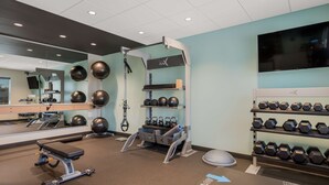 A gym with exercise equipment, a mirror, a TV, and a wall-mounted shelf with dumbbells. - Tru by Hilton Greensboro, NC (Greensboro)