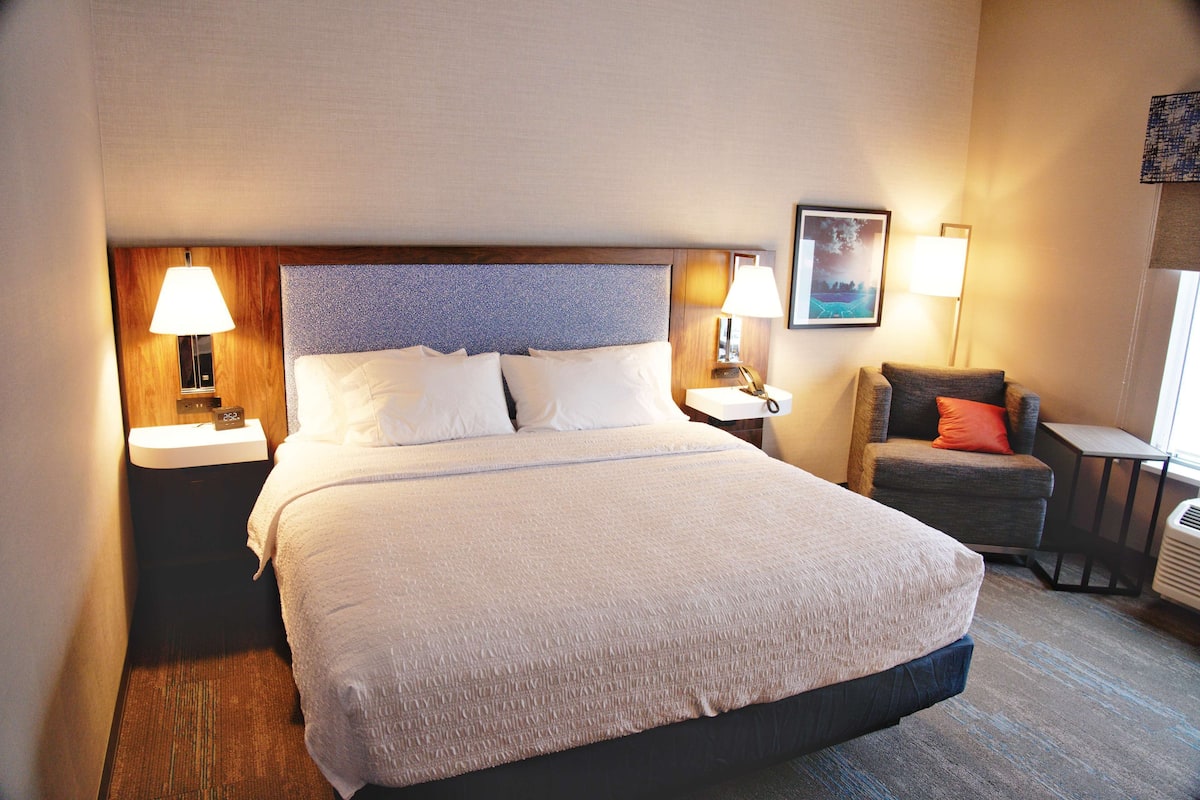 Une chambre d’hôtel avec un grand lit, deux tables de chevet avec des lampes, un petit fauteuil et un tableau au mur.