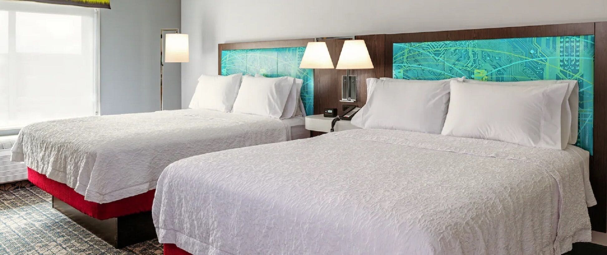Habitación de hotel con dos camas, una mesita de noche, una lámpara y un muro con diseño de circuito electrónico.