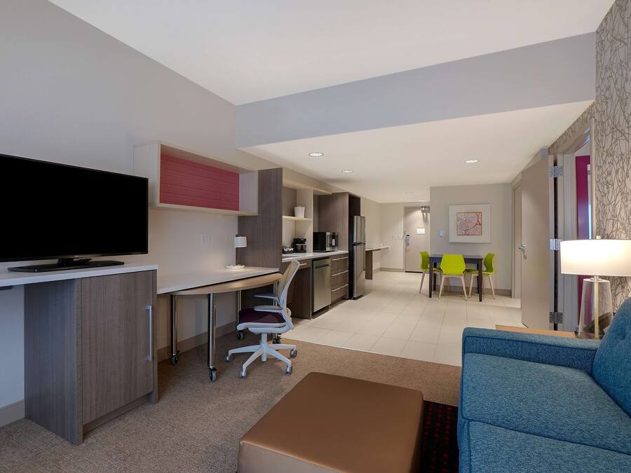 A modern hotel room with a flat-screen TV, a desk, a sofa, and a dining area with chairs.
