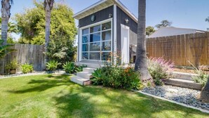 A modern house with a large window, a wooden deck, and a well-maintained garden with various plants and a palm tree. - NEW Super Clean 3 Bed / 2 Bath Home - Fully Loaded (San Diego)