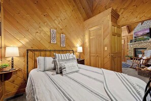 A cozy log cabin bedroom with a bed, two bedside lamps, a wooden wardrobe, and a stone fireplace. - Location! Smokies Log Cabin, Hot Tub, Fireplace (Pigeon Forge)