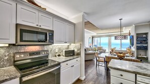 Eine moderne Küche mit integriertem Mikrowellenherd, Backofen und Toaster. Weiße Schränke, eine Marmorarbeitsplatte und ein Blick auf den Ozean durch große Fenster.