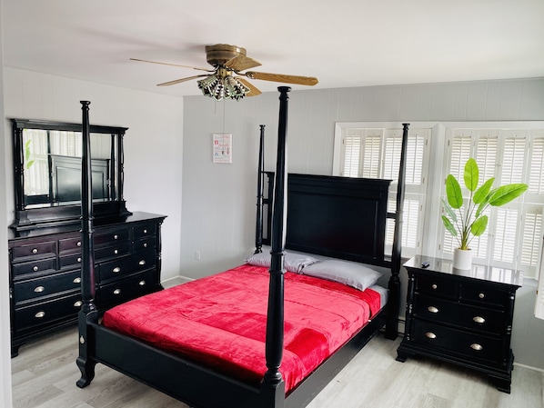 A four-poster bed with a red bedspread, a ceiling fan, a mirror, and a potted plant by the window. - Elegant and Bright Ocean Block Home w/ Private Balcony, BBQ & Patio (Seaside Park)