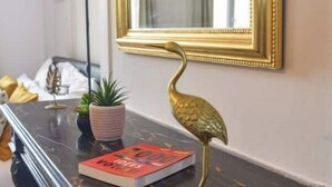 Ein Zimmer mit Schreibtisch aus Marmor, einem spiegel mit goldener Rahmung, einer dekorativen Vogelstatue, einem Buch mit dem Titel „1000 Voices“ und einer Topfpflanze.