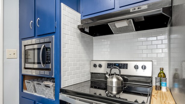 Une cuisine avec des armoires bleues, une cuisinière en acier inoxydable, un four à micro-ondes et une étagère en bois où sont rangés des ustensiles de cuisine.