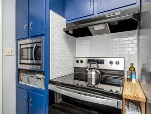 Une cuisine avec des armoires bleues, une cuisinière en acier inoxydable, un four à micro-ondes et une étagère en bois où sont rangés des ustensiles de cuisine.