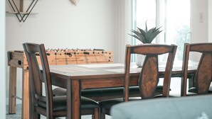 Un coin repas avec une table en bois, quatre chaises et une table de baby-foot. On trouve une plante sur la table et une fenêtre qui laisse entrer la lumière naturelle.