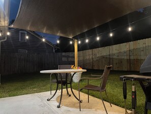 Un patio exterior con una mesa, sillas, una parrilla y luces colgantes.