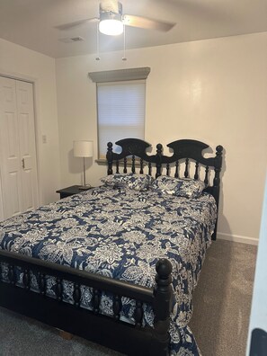 Un dormitorio con cama, mesita de noche, lámpara, ventana con persianas, ventilador de techo y puerta.