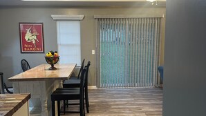 A dining area with a wooden table, chairs, and a fruit bowl. There is a ceiling light and a window with blinds.