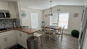 A compact kitchen with white cabinets, a microwave, and a sink. There is a dining area with a table and chairs, and a small trash bin.
