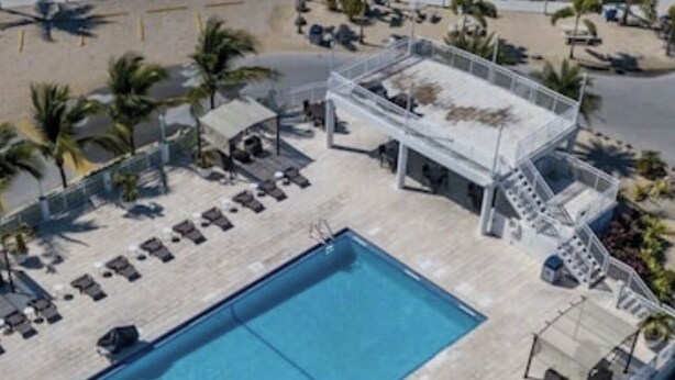 An aerial view of a resort with a large rectangular pool surrounded by sun loungers and a covered area with stairs leading to a deck.