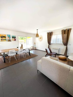 A spacious living area with a dining table, chairs, a sofa, and a large window with curtains.
