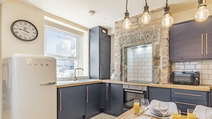 A modern kitchen with dark cabinets, a white refrigerator, a stone accent wall, and a dining area set for breakfast. - 2 bedroom accommodation in Bakewell (Bakewell)