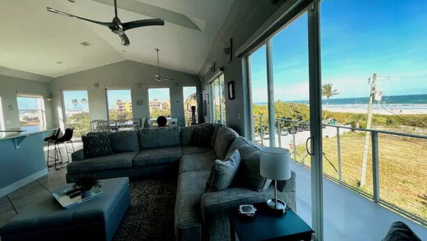 Una sala de estar moderna con un sofá seccional grande, una mesa de centro y un comedor. Varias ventanas ofrecen vista a la playa y al océano.