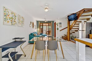 A modern living room with a glass dining table, teal sofa, and a kitchen area with a refrigerator and wooden staircase.