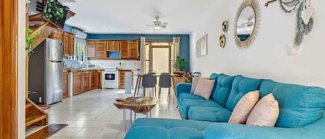 A modern living room with a blue sofa, a dining area with chairs, and a kitchen with wooden cabinets and a refrigerator.