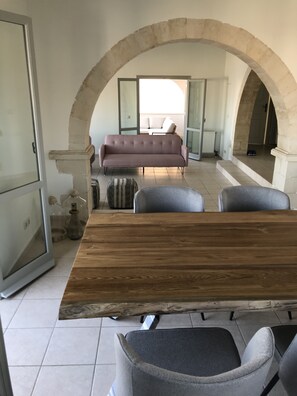 Ein Essbereich mit Holztisch und grauen Stühlen, einer gewölbten Steinarchitektur, einer Couch und Blick ins Freie.