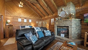 A cozy log cabin interior with a stone fireplace, leather sofa, and wooden beams.