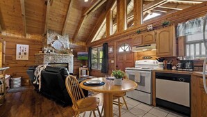 A cozy cabin interior with a fireplace, wooden walls, a dining area, and a kitchen.