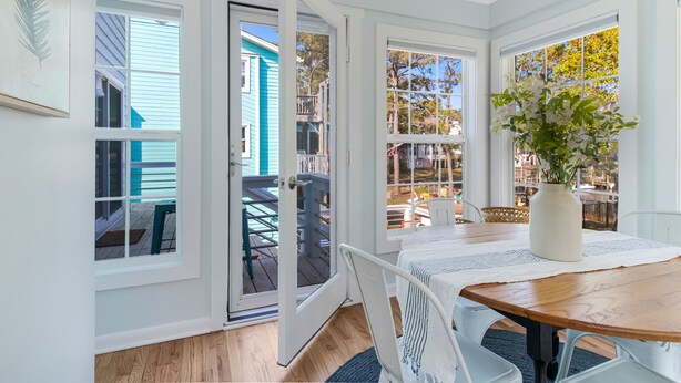 Une pièce lumineuse aux murs blancs, avec une table en bois, de grandes fenêtres et une vue sur une maison bleue à l’extérieur.
