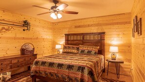 Un dormitorio acogedor con pared de troncos de madera, una cama con colcha estampada, dos lámparas de noche, un aparador de madera y un ventilador de techo.