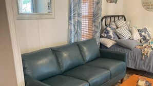 Un salon avec un canapé en cuir bleu, une table basse en bois sur laquelle reposent des dessous de verre en forme de tortue, un tapis à motifs, un lit recouvert d’une couvre-lit à fleurs bleues et blanches, un miroir et une horloge murale.