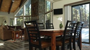 Un comedor con mesa y sillas de madera, chimenea de piedra y ventanas amplias con vistas al exterior.