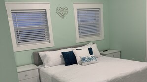 A bedroom with a bed, two bedside tables, a nightstand, a patterned rug, and two windows with blinds.