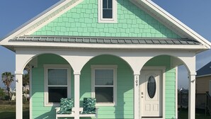 A single-story house with a green roof and white trim, featuring a front porch with a bench and a small window above the door.
