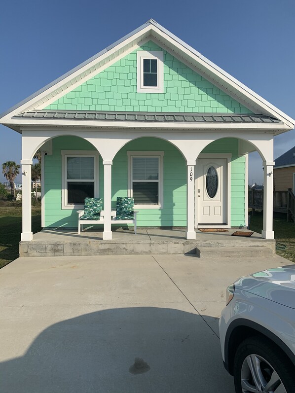 A single-story house with a green roof and white trim, a front porch with a bench, and a mailbox.