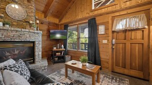 A cozy log cabin interior with a stone fireplace, a television, a wooden door, and a comfortable seating area. - Location! Cute Studio Log Cabin,Fireplace, Hot Tub (Pigeon Forge)